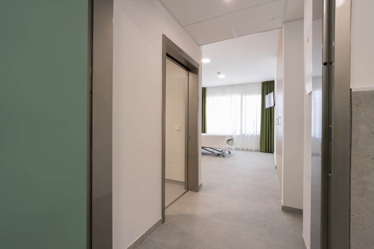 Pasillo en un centro médico; pisos gris claro, paredes blancas, puerta verde y habitación con una cama visible.