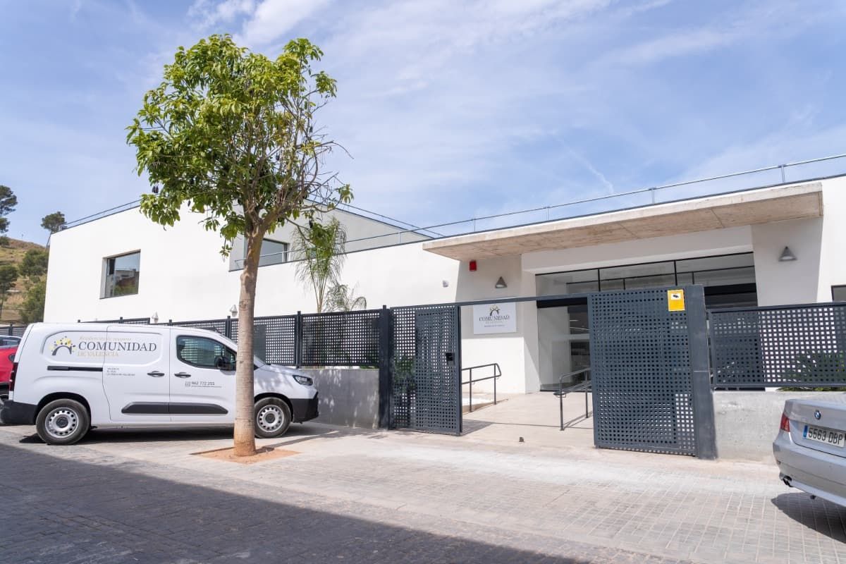 Vista exterior de un edificio blanco moderno con una valla gris oscuro, una furgoneta y un coche aparcado. Hay un árbol delante.