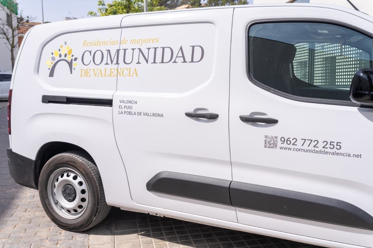 Furgoneta blanca con el logo de la Comunidad Valenciana, número de teléfono y dirección. Aparcada en la calle, con sol.