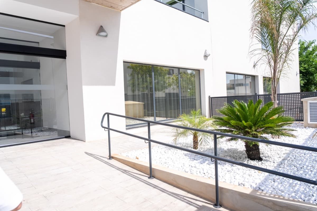 Exterior de edificio blanco con rampa, barandilla, jardinería y entrada de vidrio.