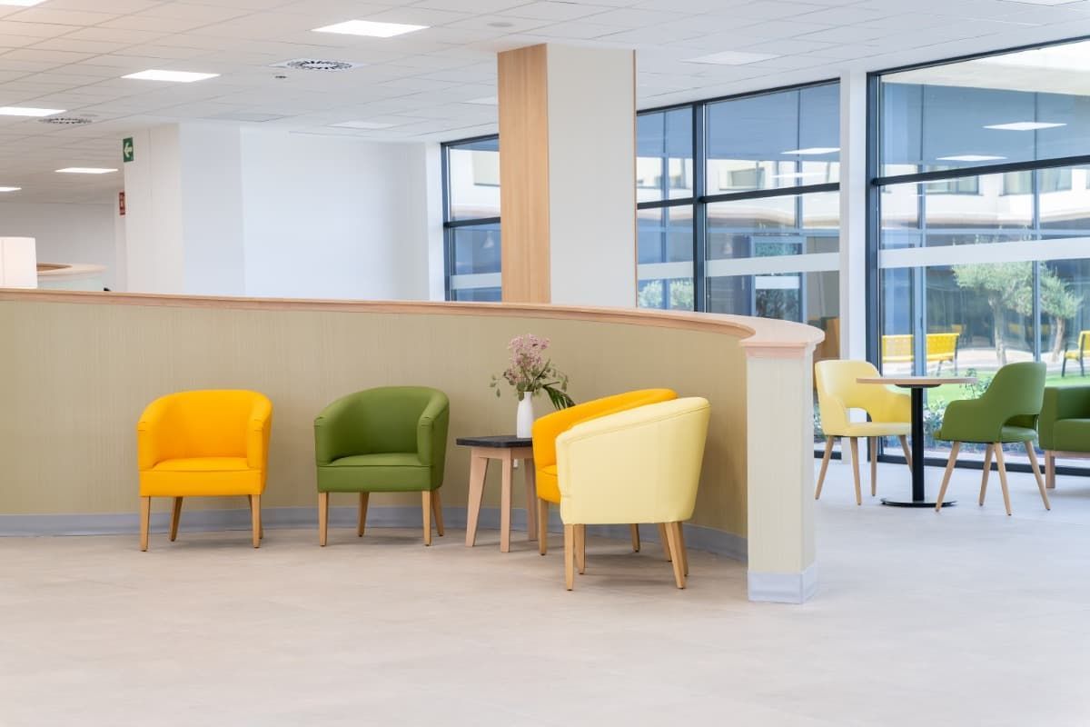 Sillas de colores en una zona de espera llena de luz, con grandes ventanales y una mampara baja.