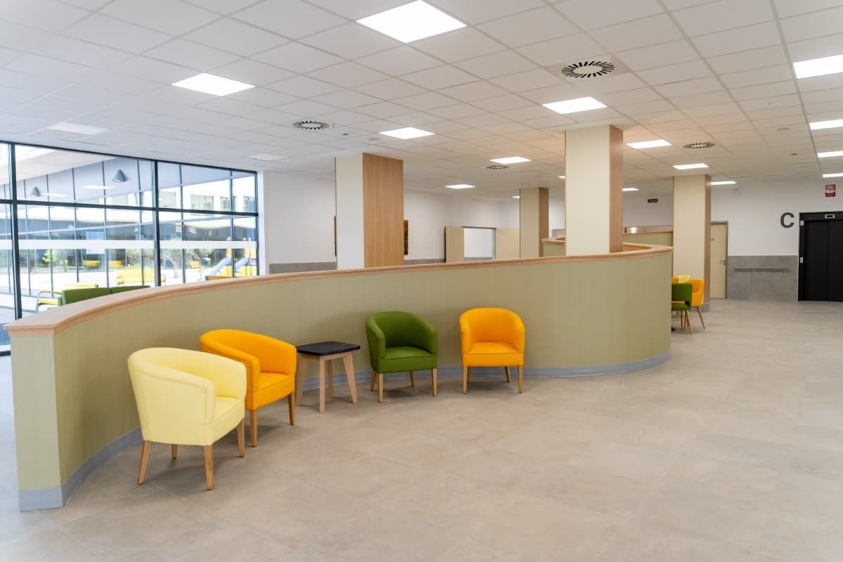 Zona de espera con sillas de colores a lo largo de una pared curva, ventanas al fondo.