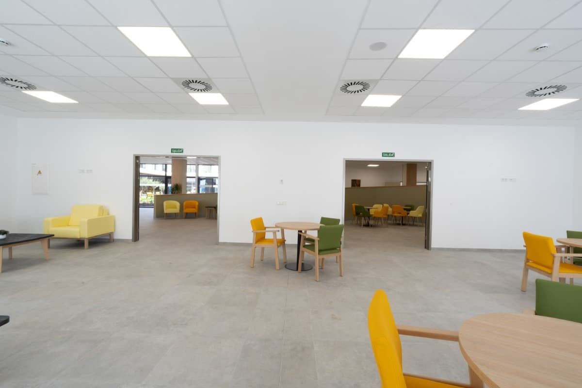 Habitación luminosa y abierta con dos puertas que dan a zonas con mesas y sillas. Paredes blancas, suelo gris y sillas amarillas y verdes.