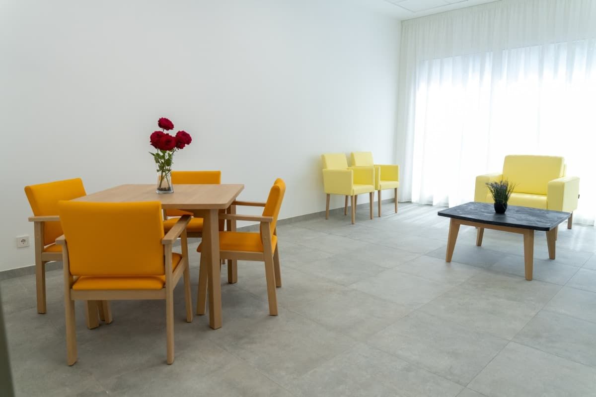 Una sala de estar moderna y luminosa: sillas amarillas, una mesa cuadrada, una mesa de café oscura y un jarrón con flores rojas.