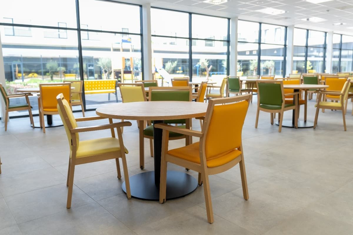 Comedor luminoso con mesas redondas, sillas de varios colores y grandes ventanales.