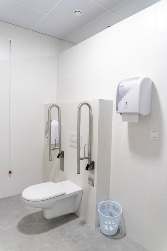Baño accesible con inodoro, barras de apoyo y dispensador de toallas de papel; paredes y suelo blancos.