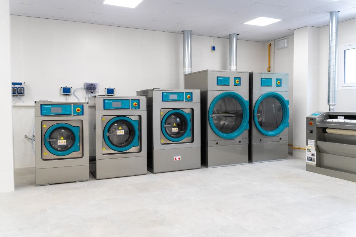Fila de lavadoras industriales grises con detalles en verde azulado en una habitación blanca.