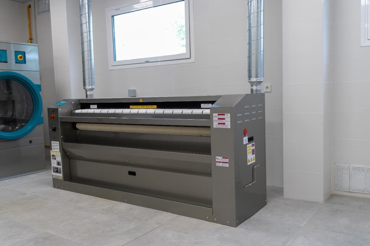 Planchadora industrial en lavadero, máquina gris con rodillo grande, ventana al fondo.