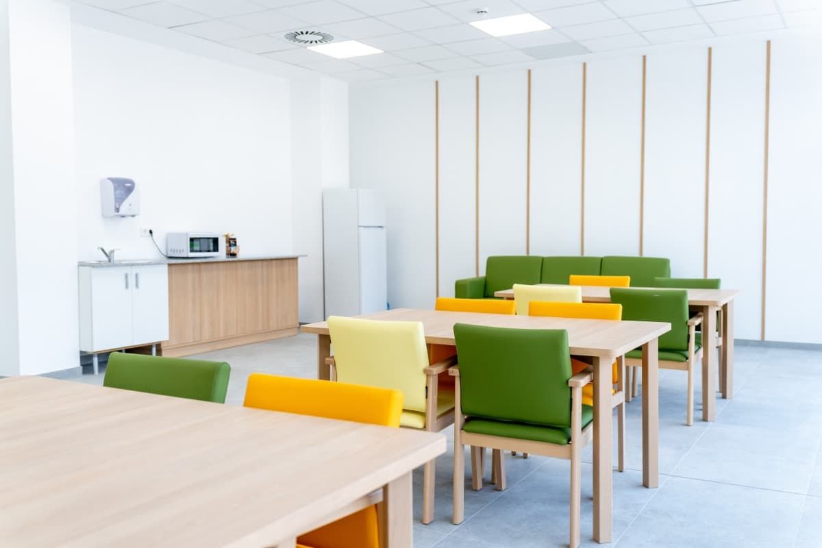 Cafetería luminosa y moderna con mesas de madera y sillas de colores; un sofá y un microondas contra una pared blanca.
