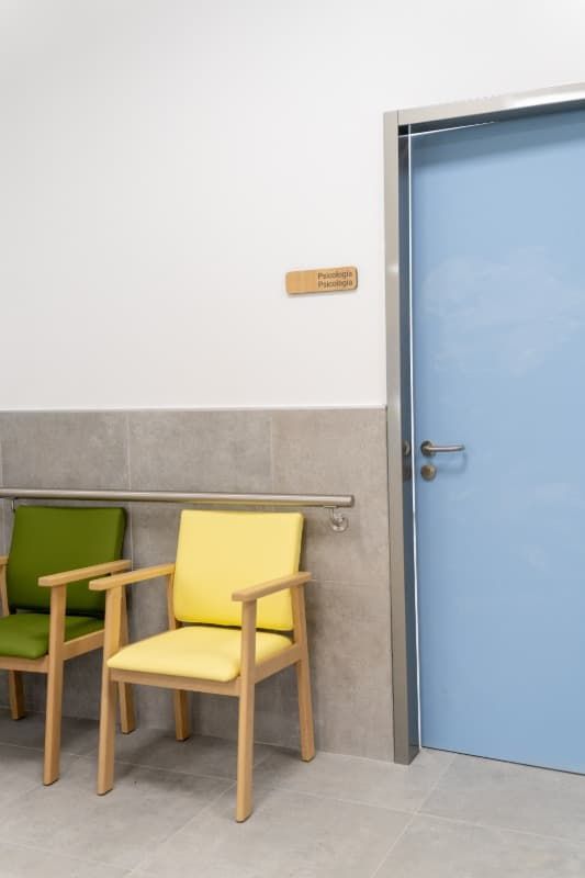 Sala de espera con sillas verdes y amarillas, puerta azul claro.