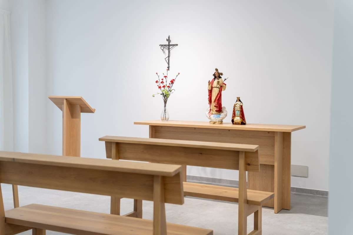 Una capilla escasamente amueblada con un altar de madera, bancos y un atril. Un crucifijo y estatuas reposan sobre el altar.