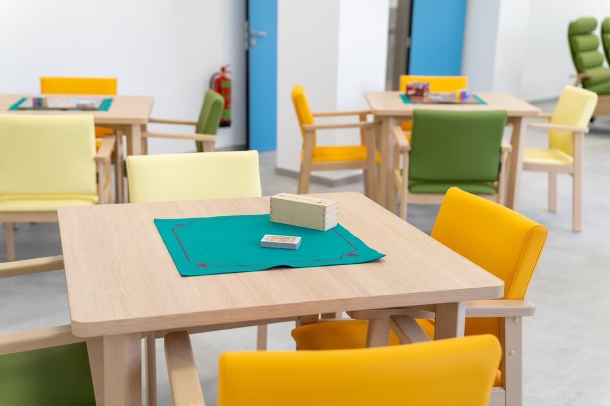 Mesas con sillas amarillas y verdes en una habitación luminosa. Una caja y cartas descansan sobre un tapete verde.