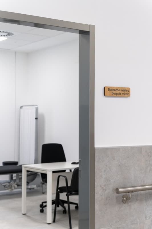 Una puerta que conduce a una oficina blanca con un escritorio, sillas y equipo médico; hay un cartel en la pared.