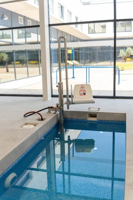 Piscina con ascensor de accesibilidad, agua azul, asiento blanco, ventanas de cristal, edificio moderno.