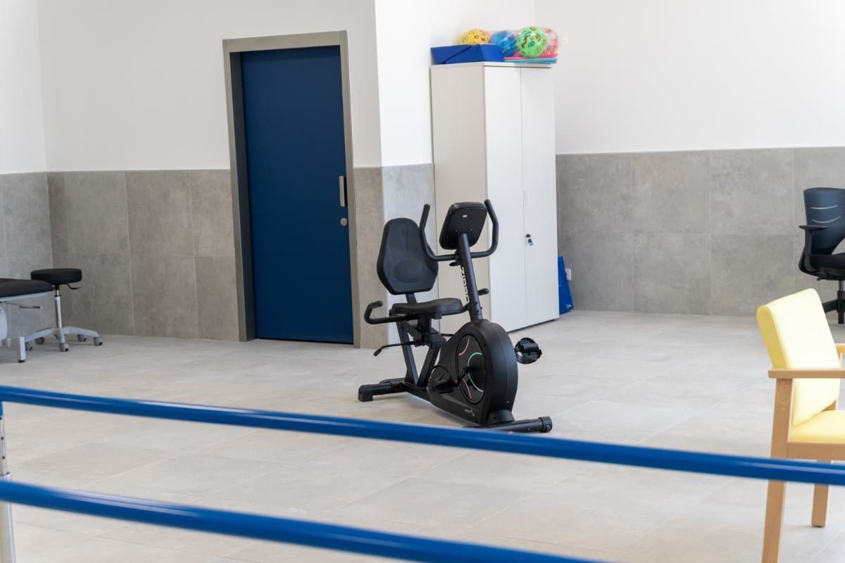 Sala de ejercicios con bicicleta reclinada, puerta azul, paredes grises y barra azul.