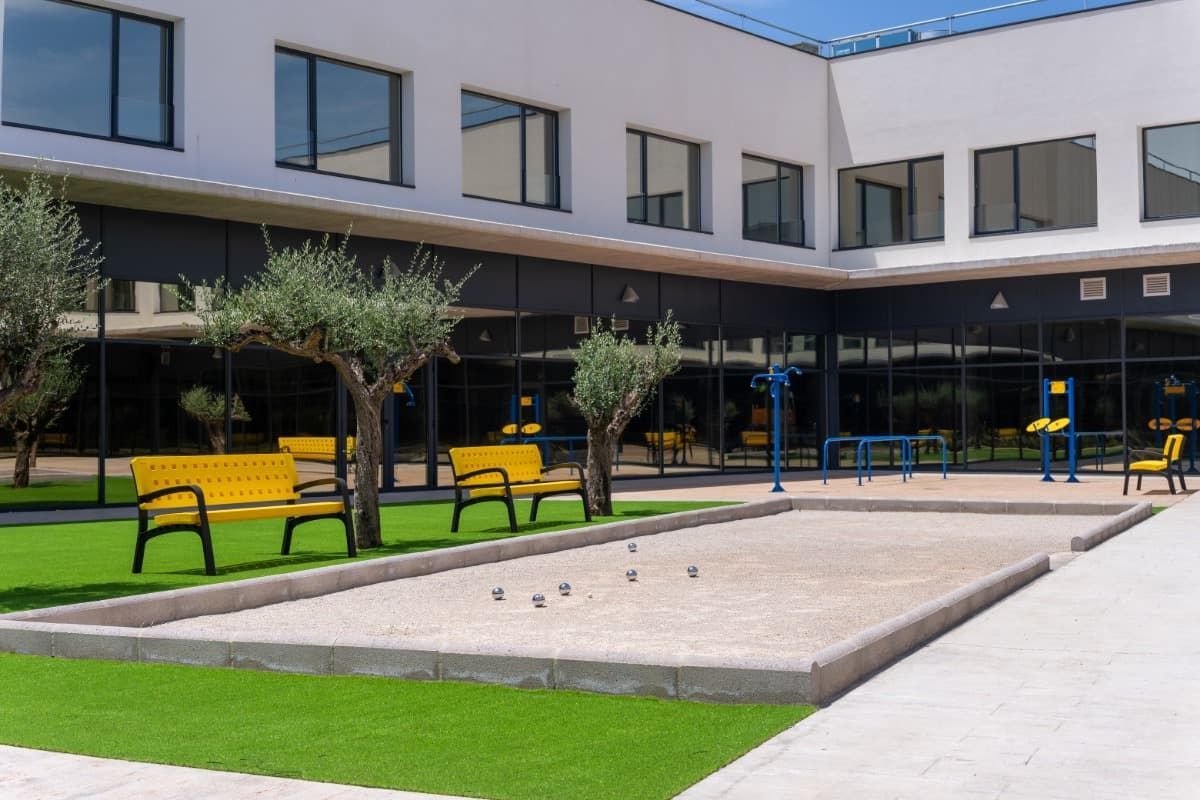 Patio con pista de petanca, bancos amarillos y árboles frente a un edificio moderno.