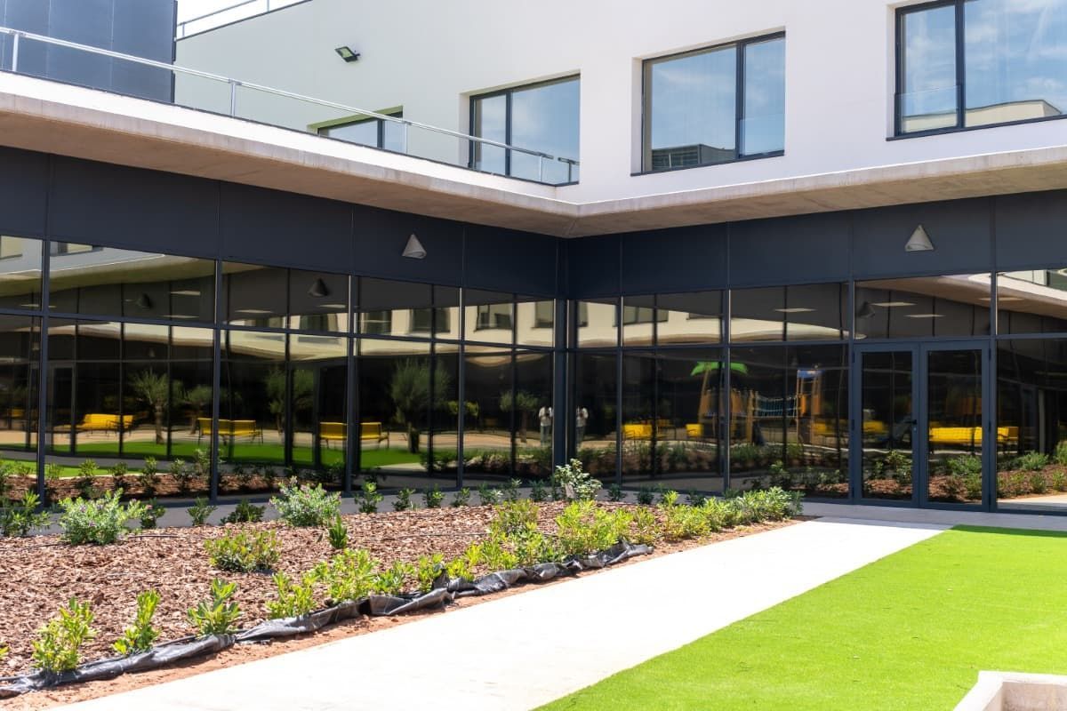 Exterior de un edificio moderno con paredes de vidrio con marco negro, un patio con césped y jardines.