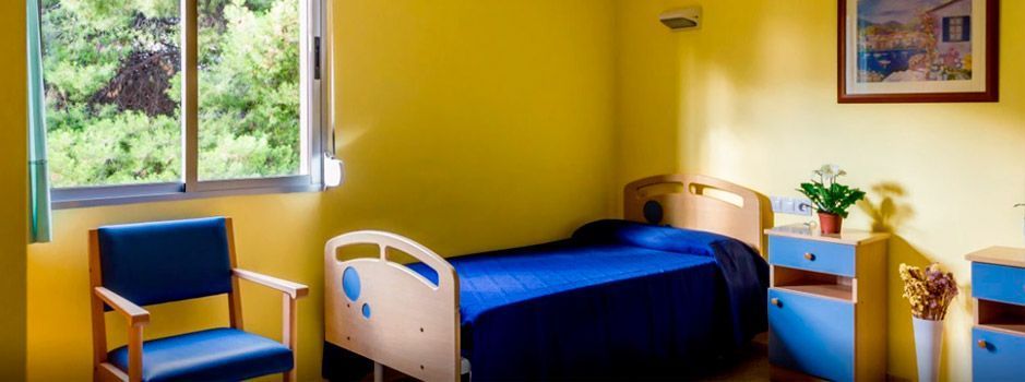 Una habitación de hospital con una cama, sillas, mesitas de noche y una ventana.