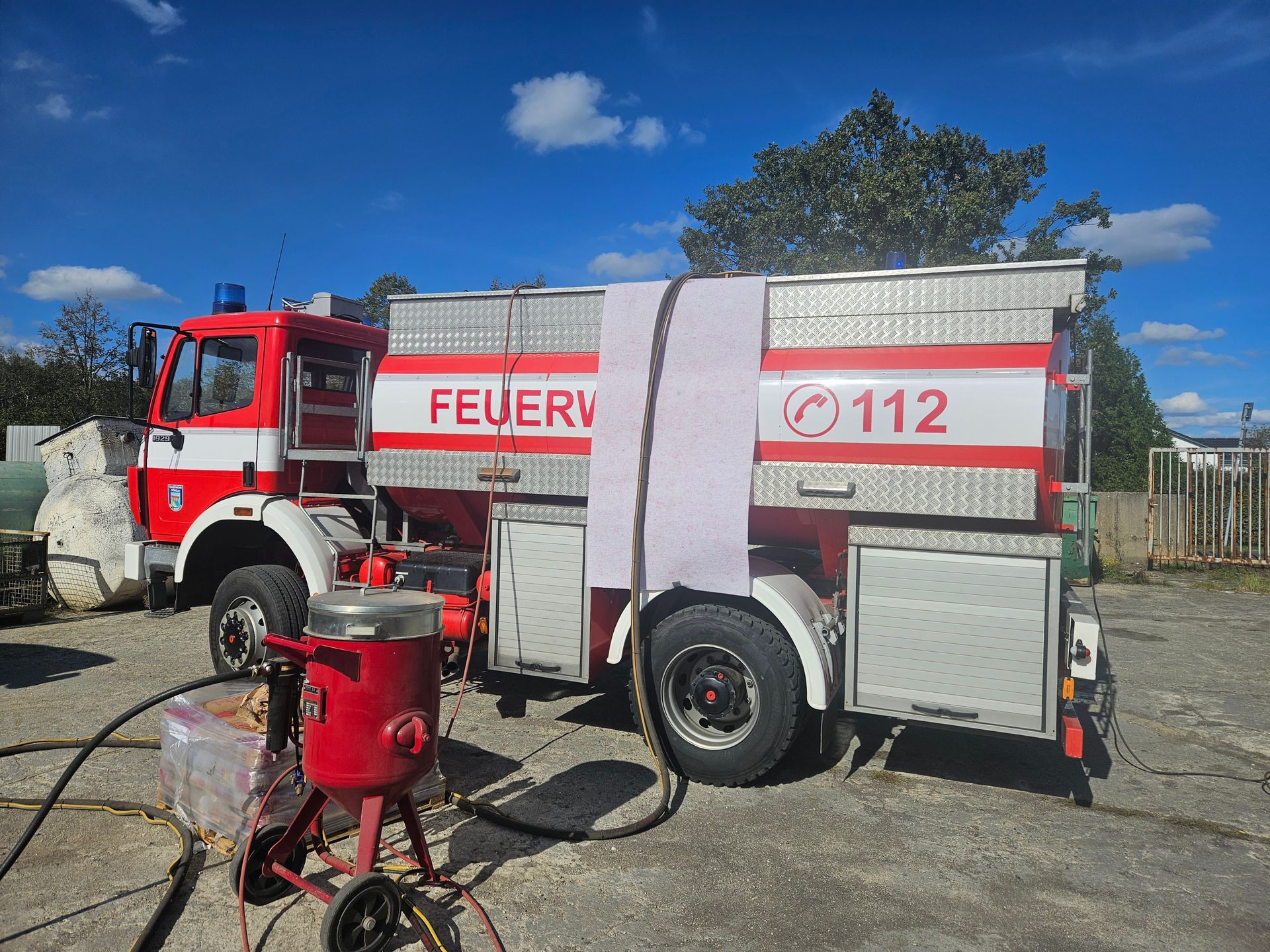 Feuerwehrauto mit rot-weißer Lackierung, geparkt auf einer gepflasterten Fläche neben einer Sandstrahlmaschine.
