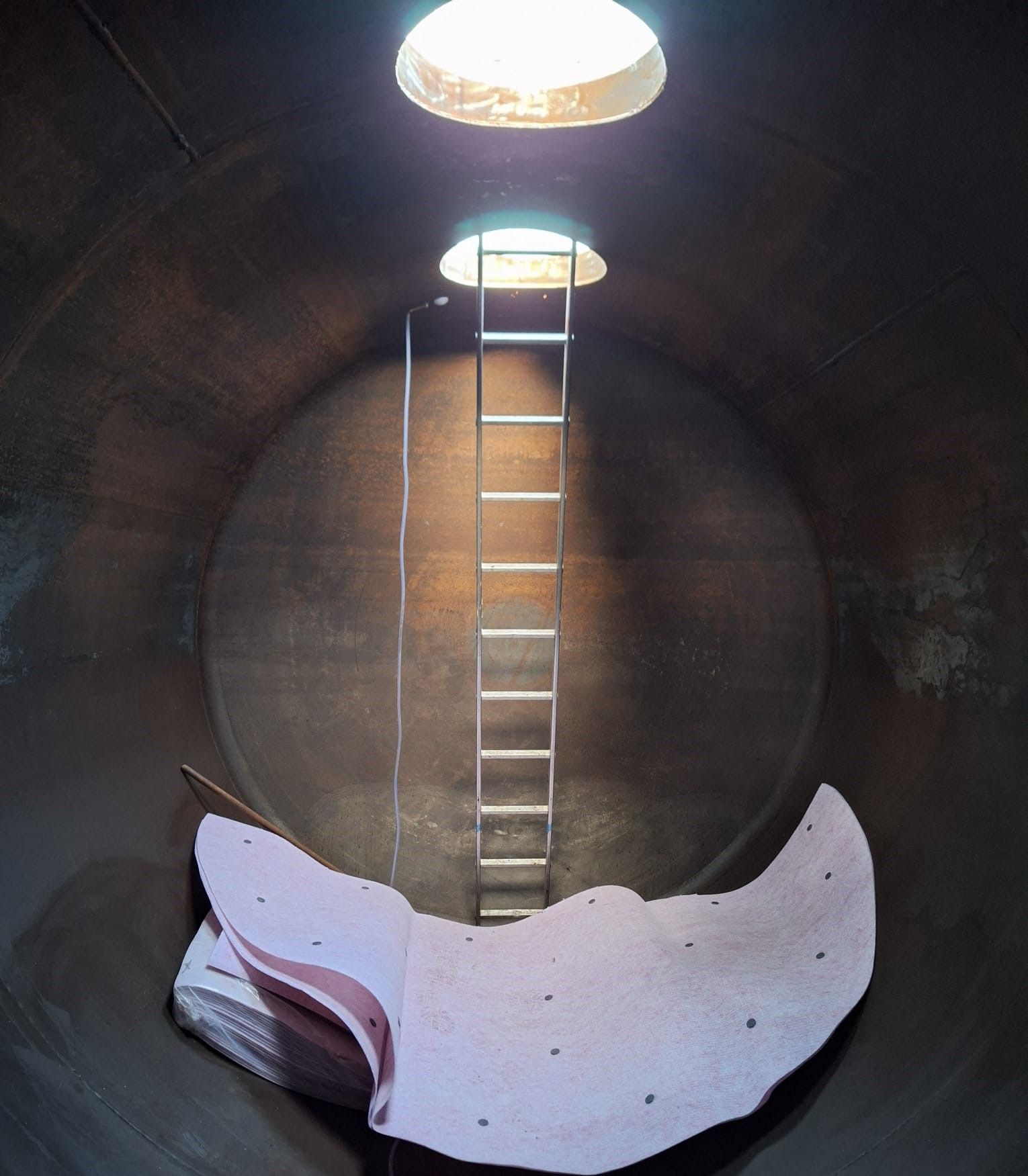 Im Inneren befindet sich ein großer, dunkler Metalltank mit einer Leiter, die zu einer runden Öffnung hinaufführt. Im unteren Teil befindet sich eine rosa Isolierung.