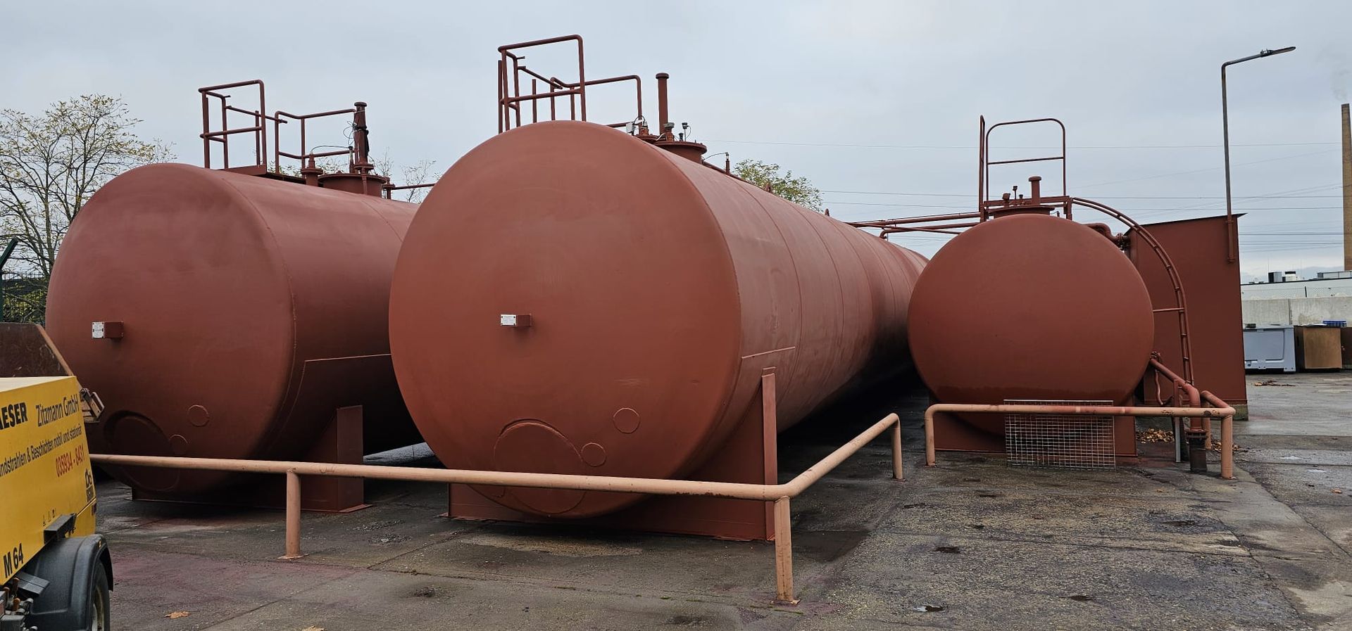 Mehrere große, rote, zylindrische Lagertanks stehen draußen auf Kies unter einem bewölkten Himmel.