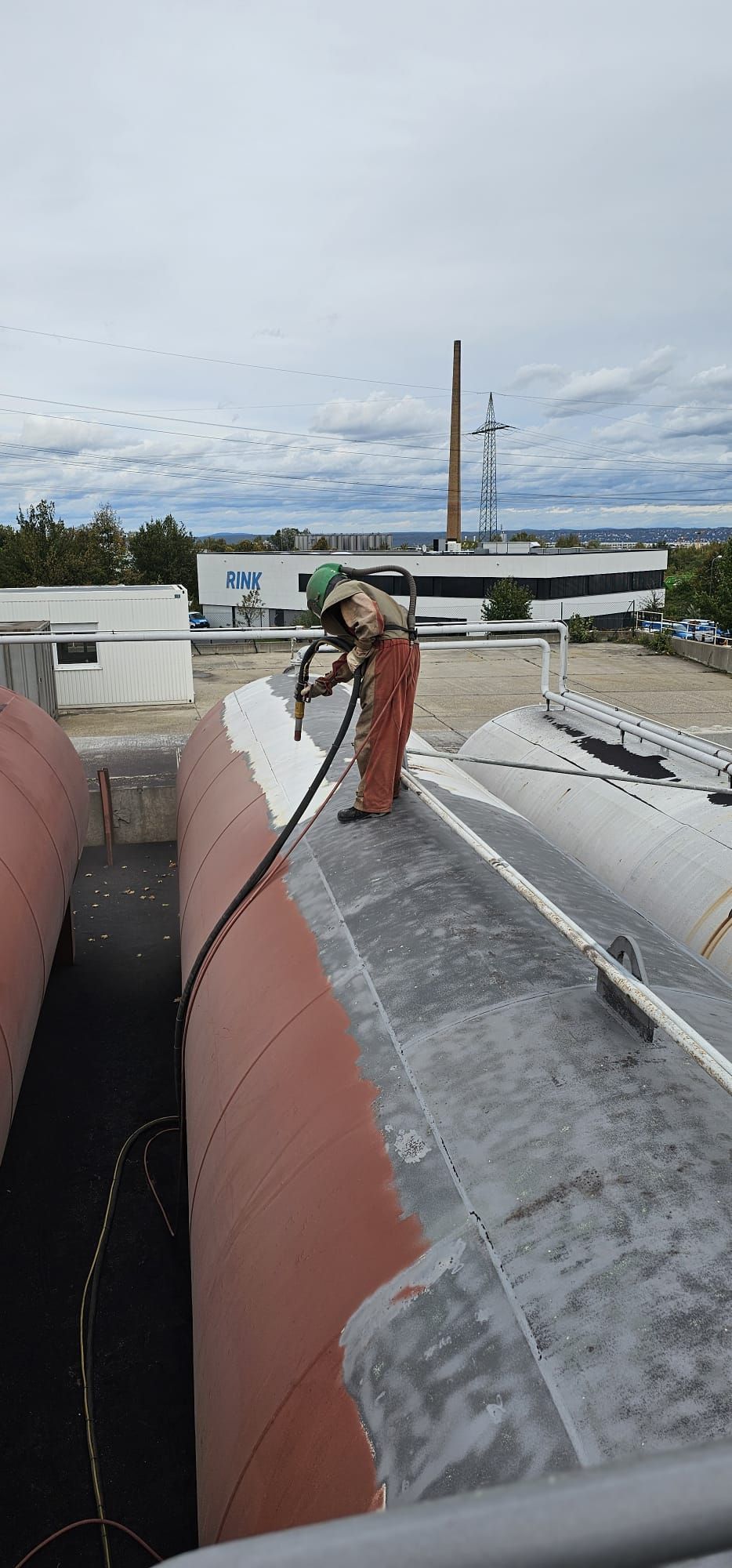 Ein Arbeiter in Sicherheitsausrüstung sandstrahlt einen großen Industrietank auf einem Dach.