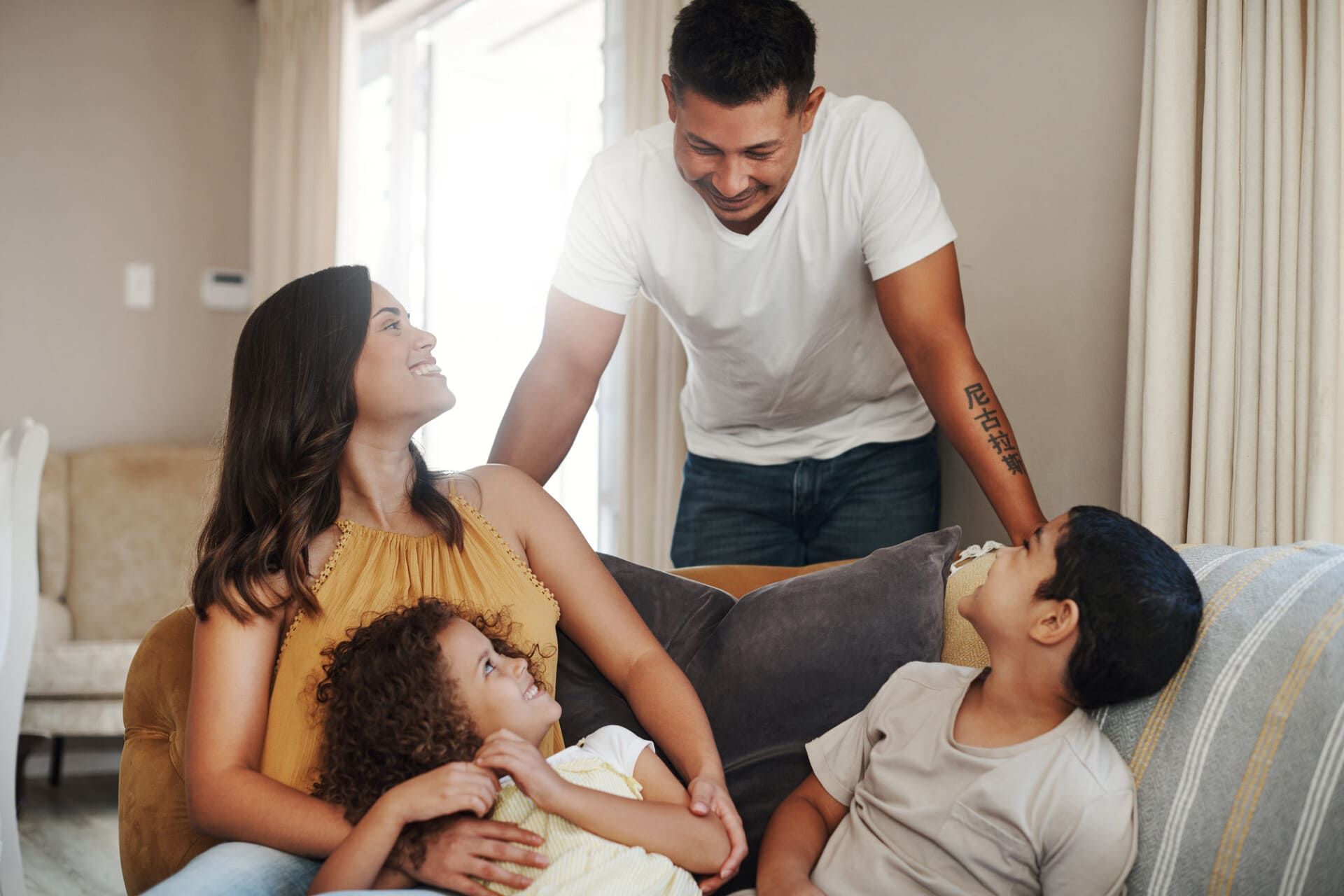 Una familia de cuatro personas se relaja en un sofá, compartiendo sonrisas e interacciones afables en una sala de estar luminosa y moderna.