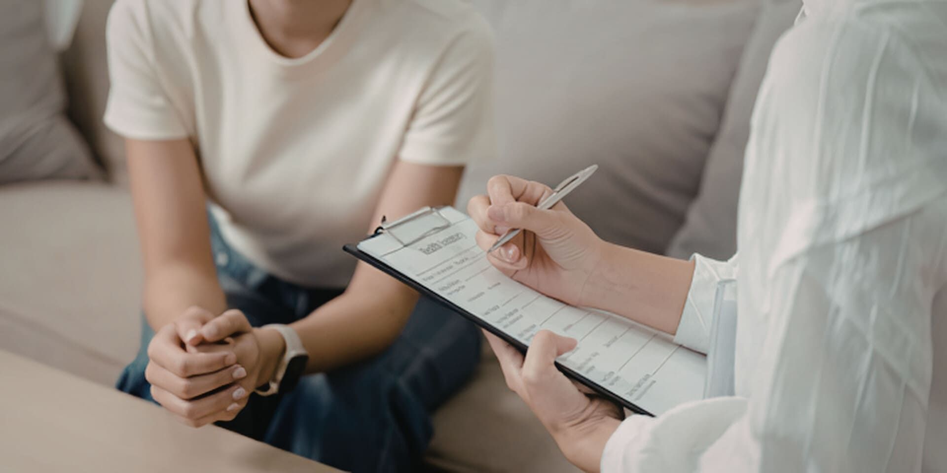 Un profesional, sentado en un sofá, toma notas en un portapapeles mientras escucha a una persona durante una sesión de terapia.