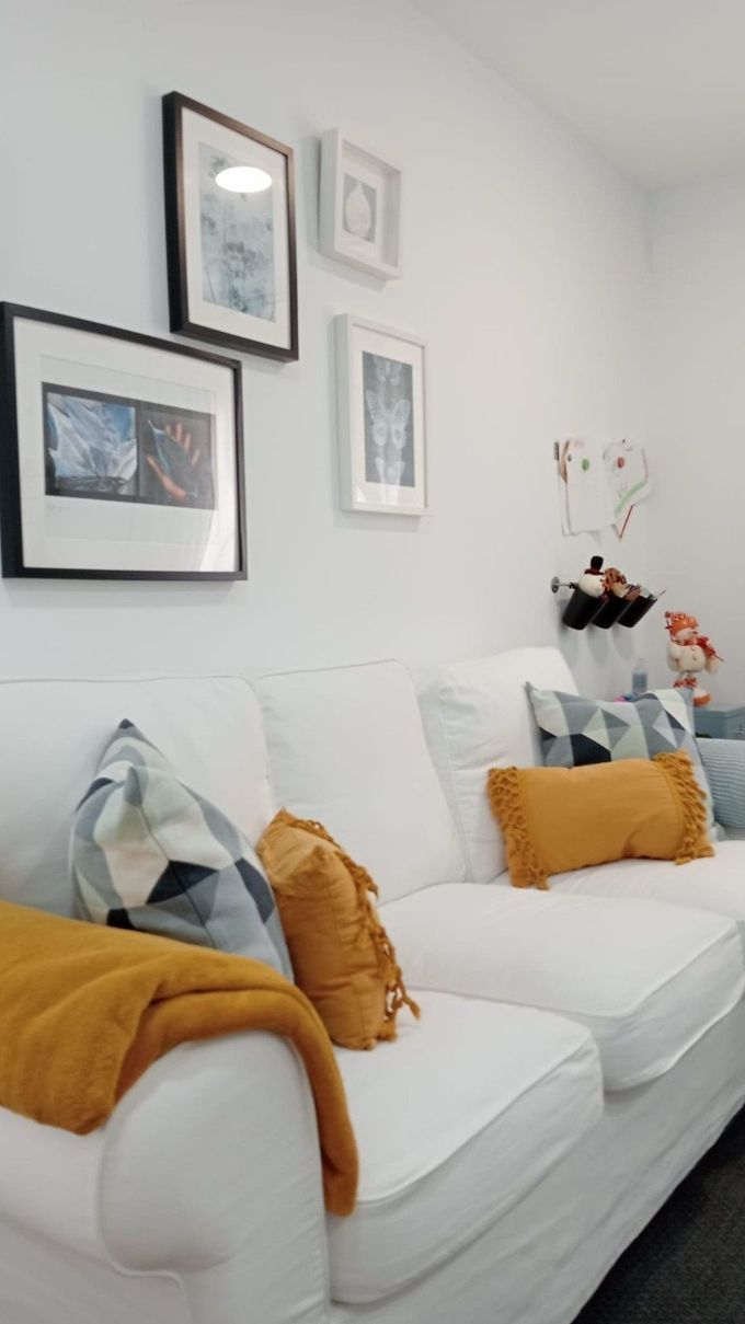 Sofá blanco con cojines de color amarillo mostaza y una manta, complementado por una pared con una galería de cuadros enmarcados.