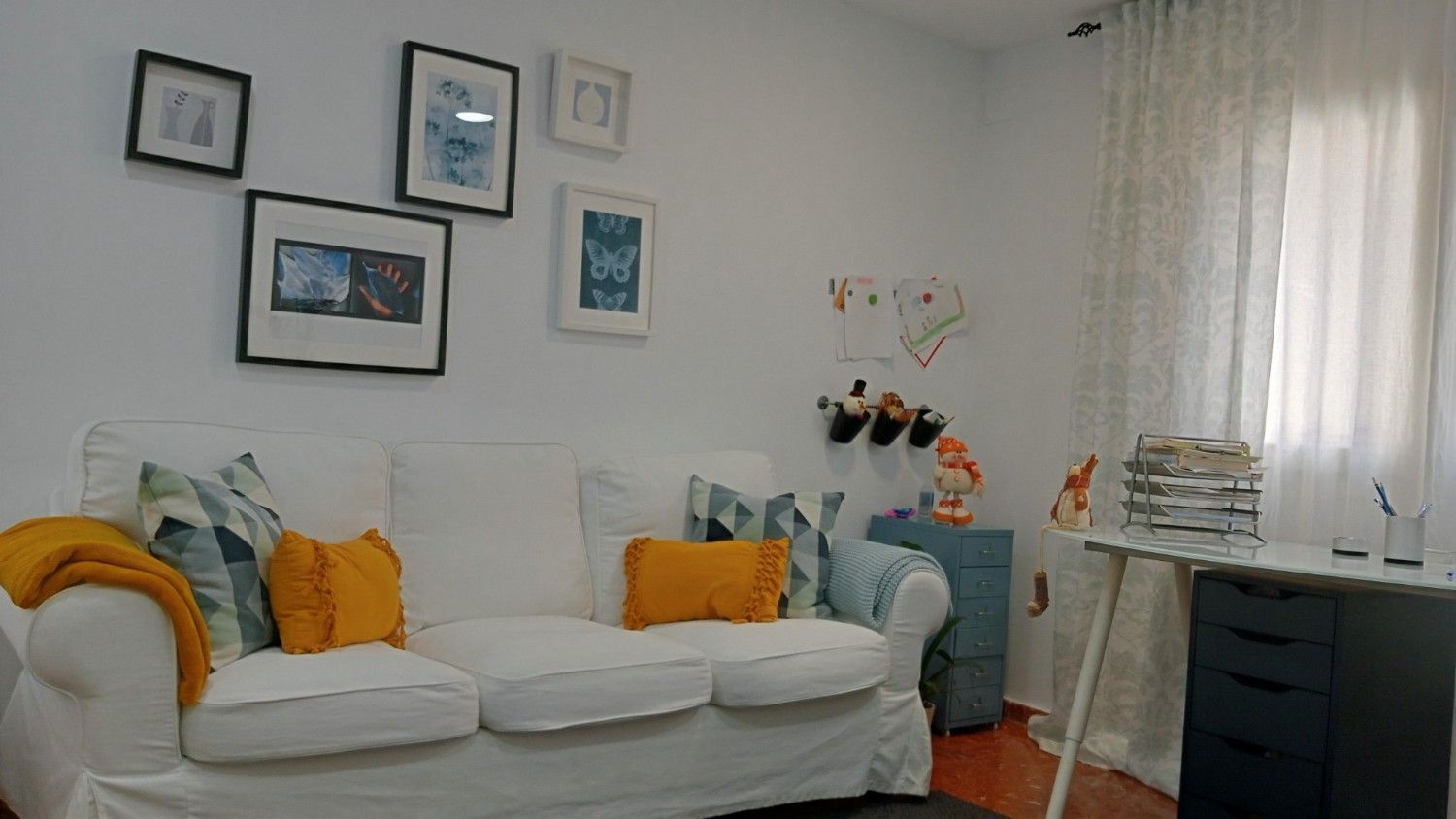 Sofá blanco con almohadas naranjas y azules en una habitación con una pared de galería, escritorio y cortinas.