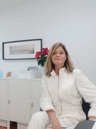 Una profesional sentada en una oficina luminosa, vestida de blanco, con un cuadro enmarcado y flores rojas sobre un armario detrás de ella.