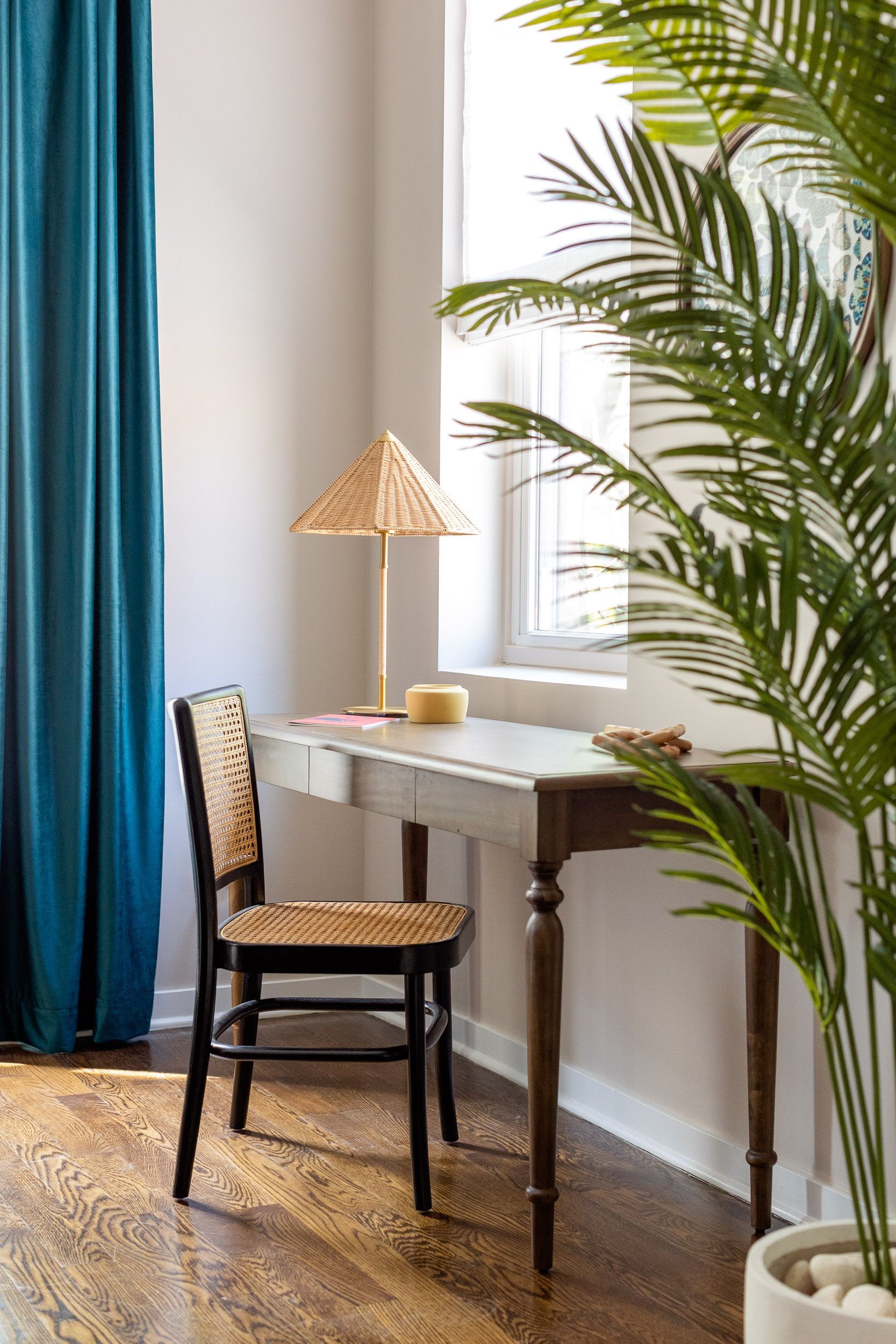 Cozy corner with woven chair, small desk lamp, potted palm, and blue curtains by a window