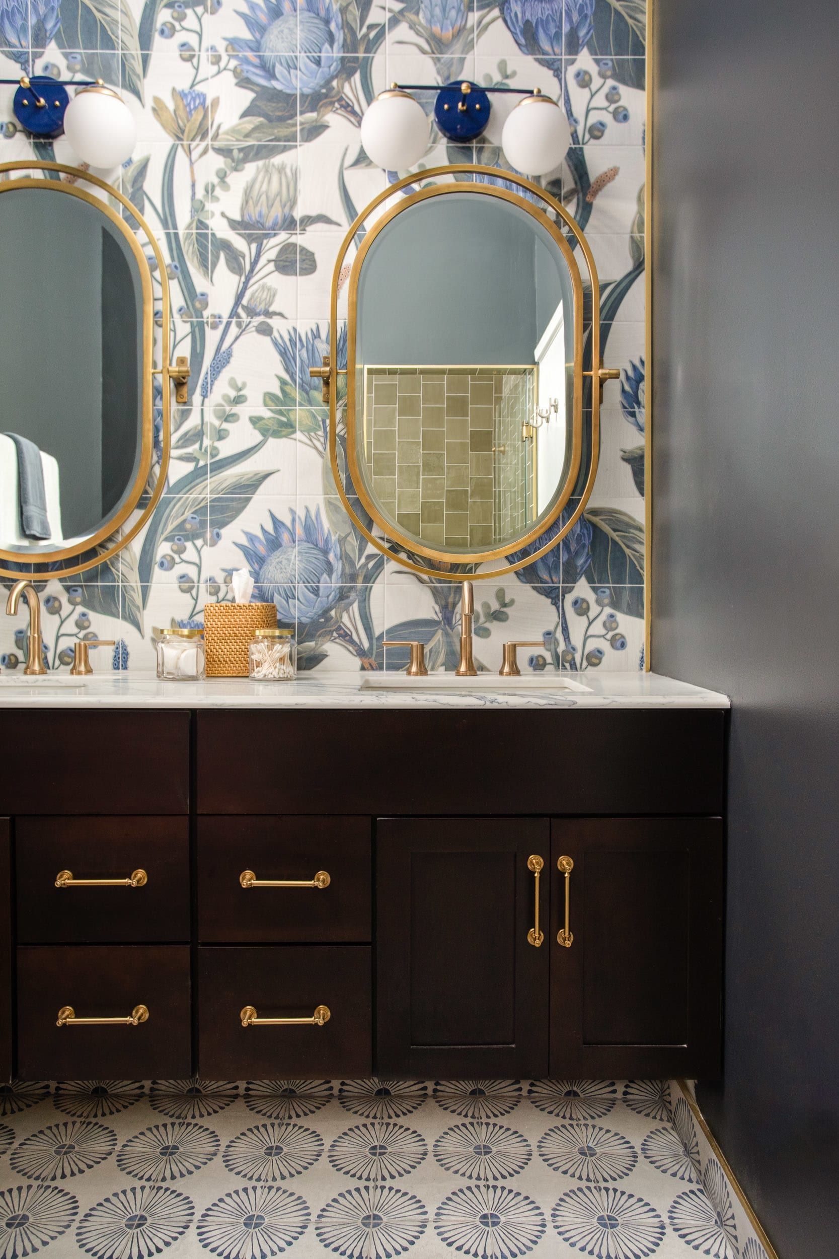 Bathroom vanity with two gold-framed oval mirrors, dark cabinets, white countertop, and blue floral wallpaper