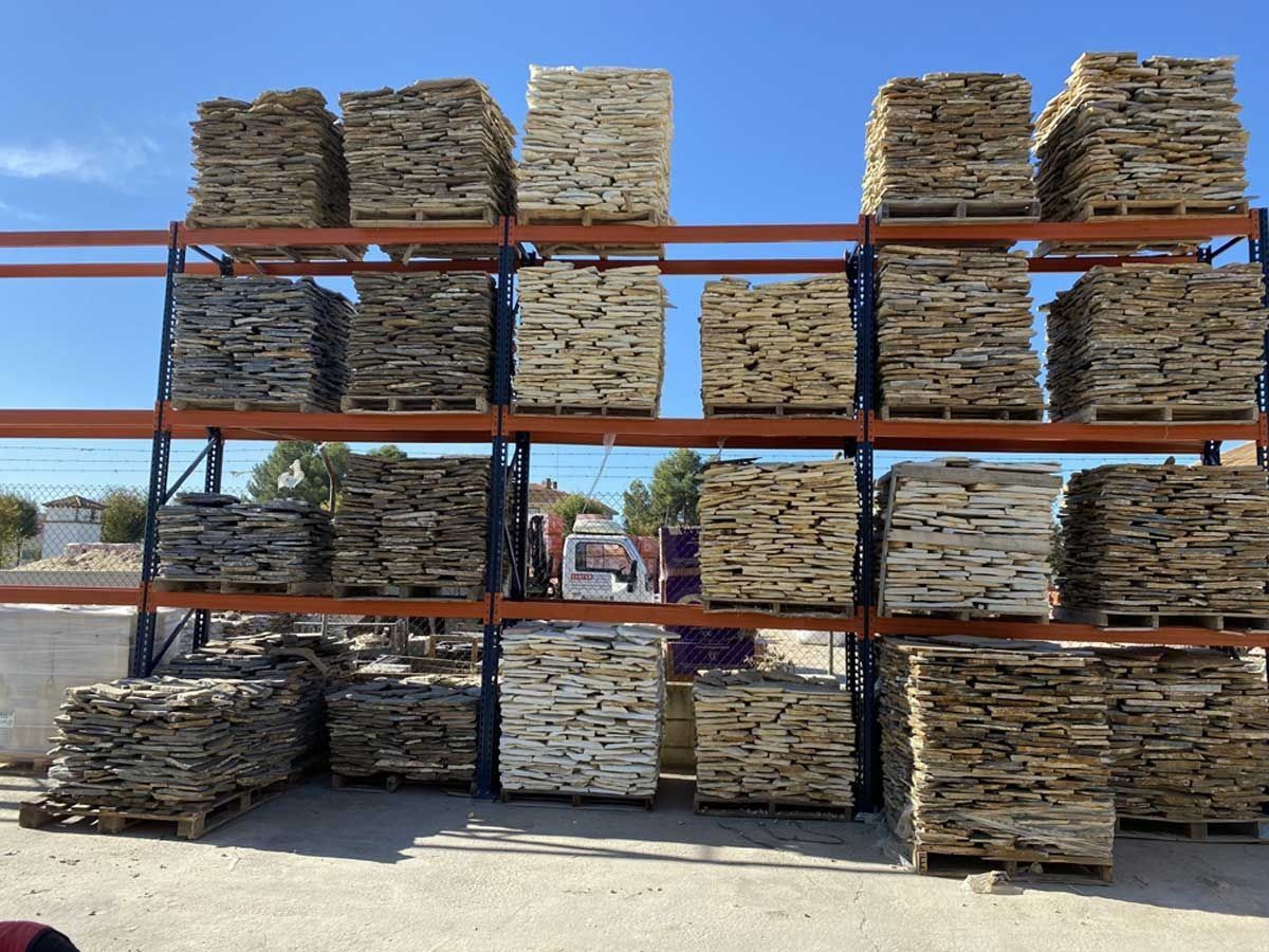 Pilas de baldosas de piedra natural de diversos tipos, dispuestas sobre estanterías metálicas de un almacén bajo un cielo azul despejado.