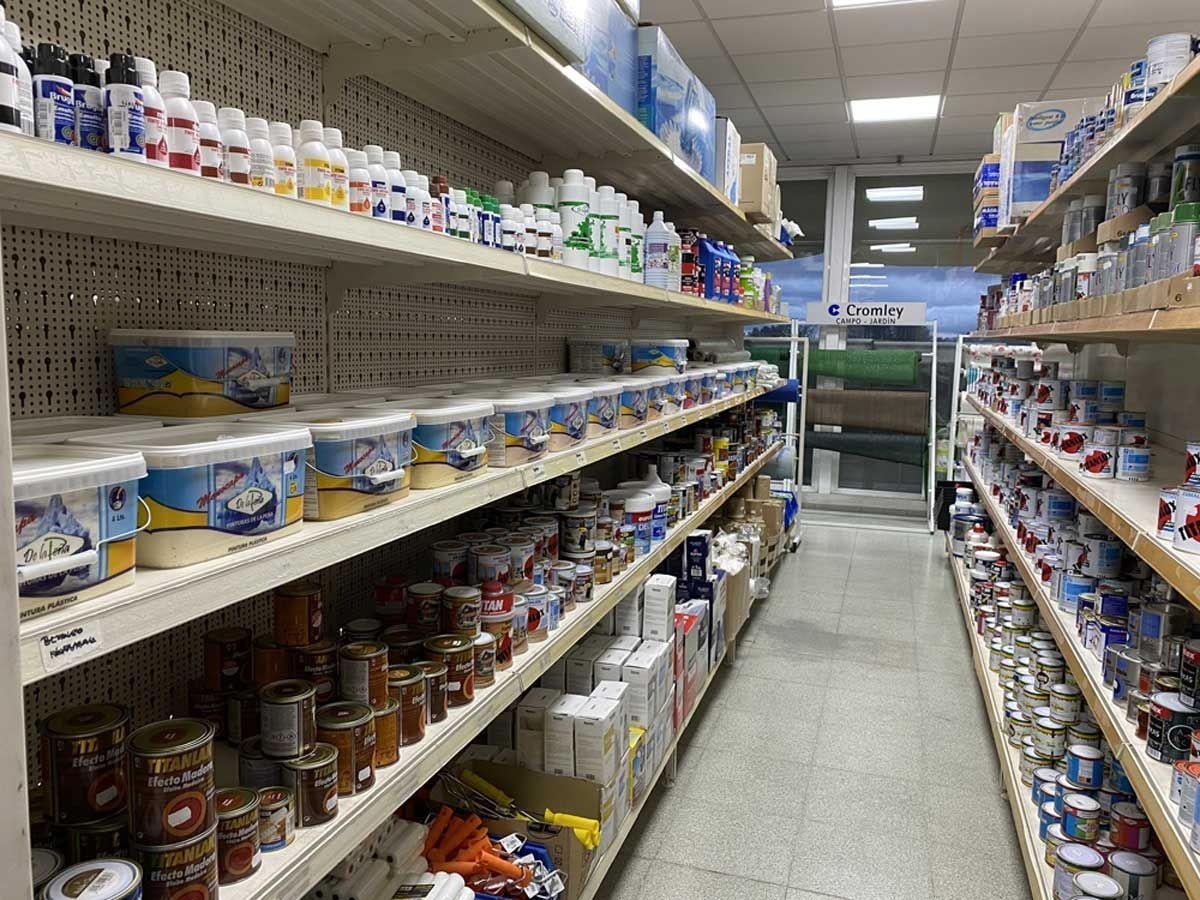 Un pasillo de una ferretería repleto de filas de latas de pintura, recipientes de plástico blanco y botellas sobre estanterías de madera.