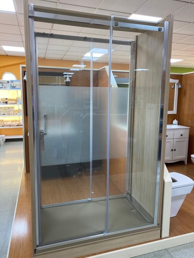 Una mampara de ducha rectangular de cristal con detalles cromados y puerta corredera esmerilada, expuesta en una sala de exhibición.
