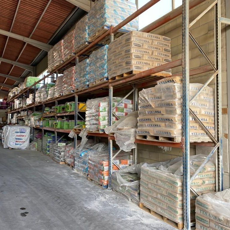 Interior de un almacén industrial con filas de altas estanterías metálicas apiladas con palés de materiales de construcción embalados.