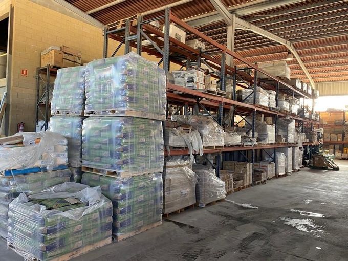 Almacén con altas estanterías metálicas que contienen múltiples palés de mercancías envueltas en plástico transparente.