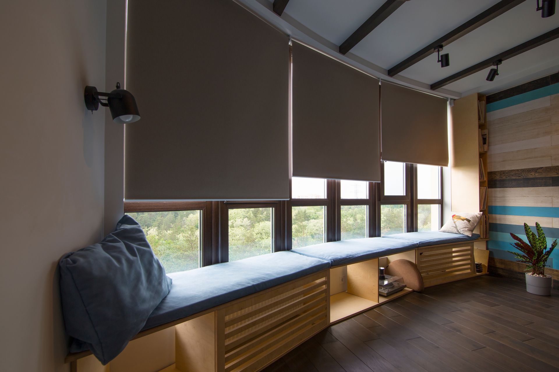 Salon moderne avec banquette rembourrée, stores gris, grandes fenêtres et plante en pot