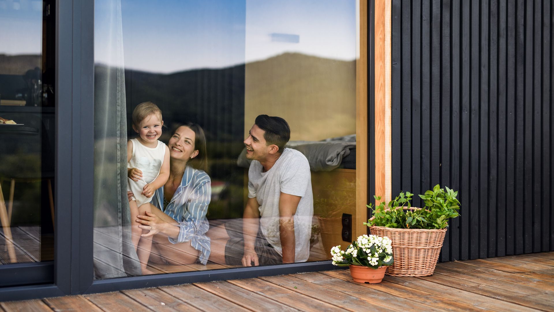 Une famille assise près d'une porte-fenêtre coulissante sur une terrasse en bois avec des fleurs en pot et des montagnes en arrière-plan