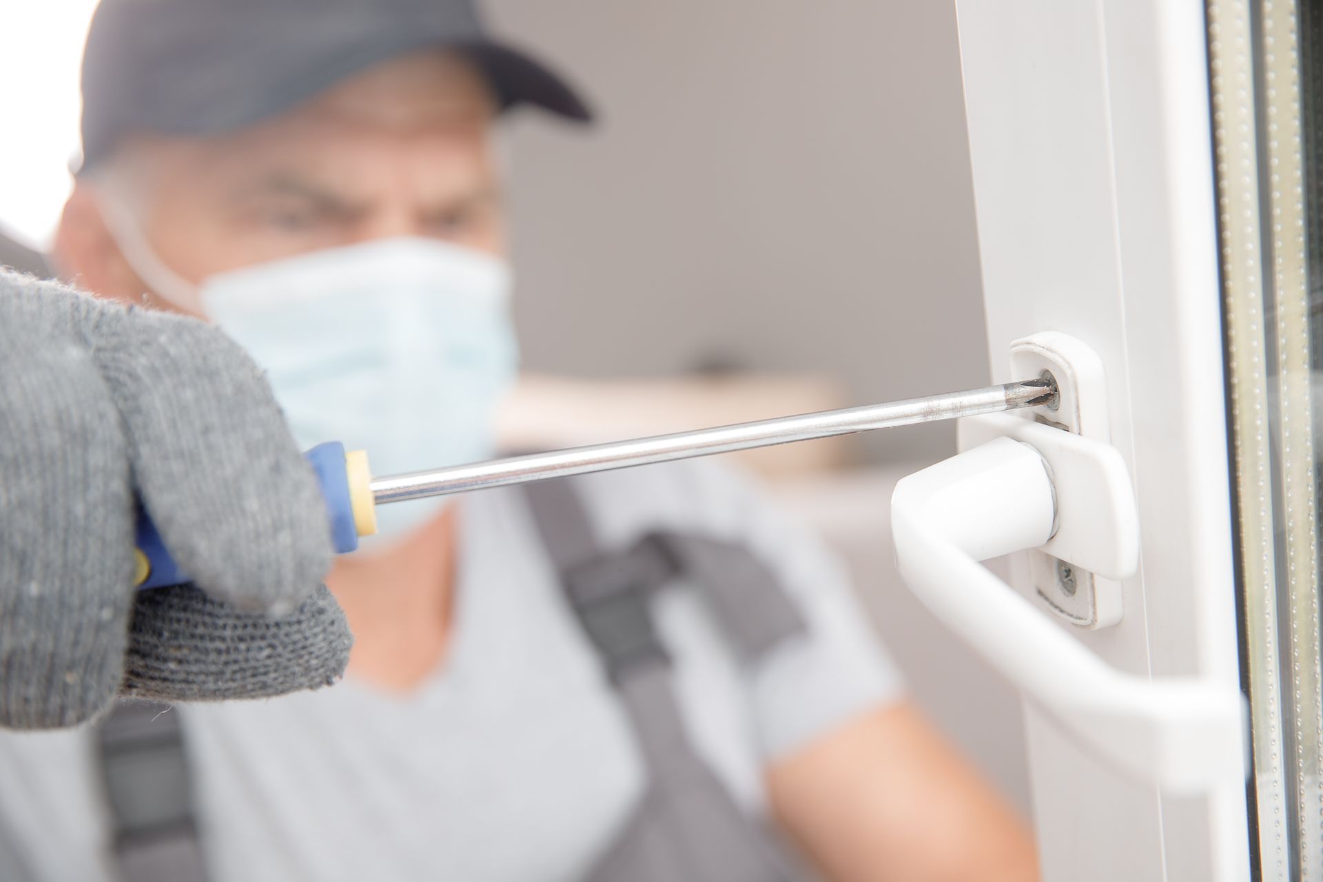 Un technicien ganté répare une poignée de fenêtre blanche à l'intérieur à l'aide d'un tournevis ; une personne masquée est floutée en arrière-plan.