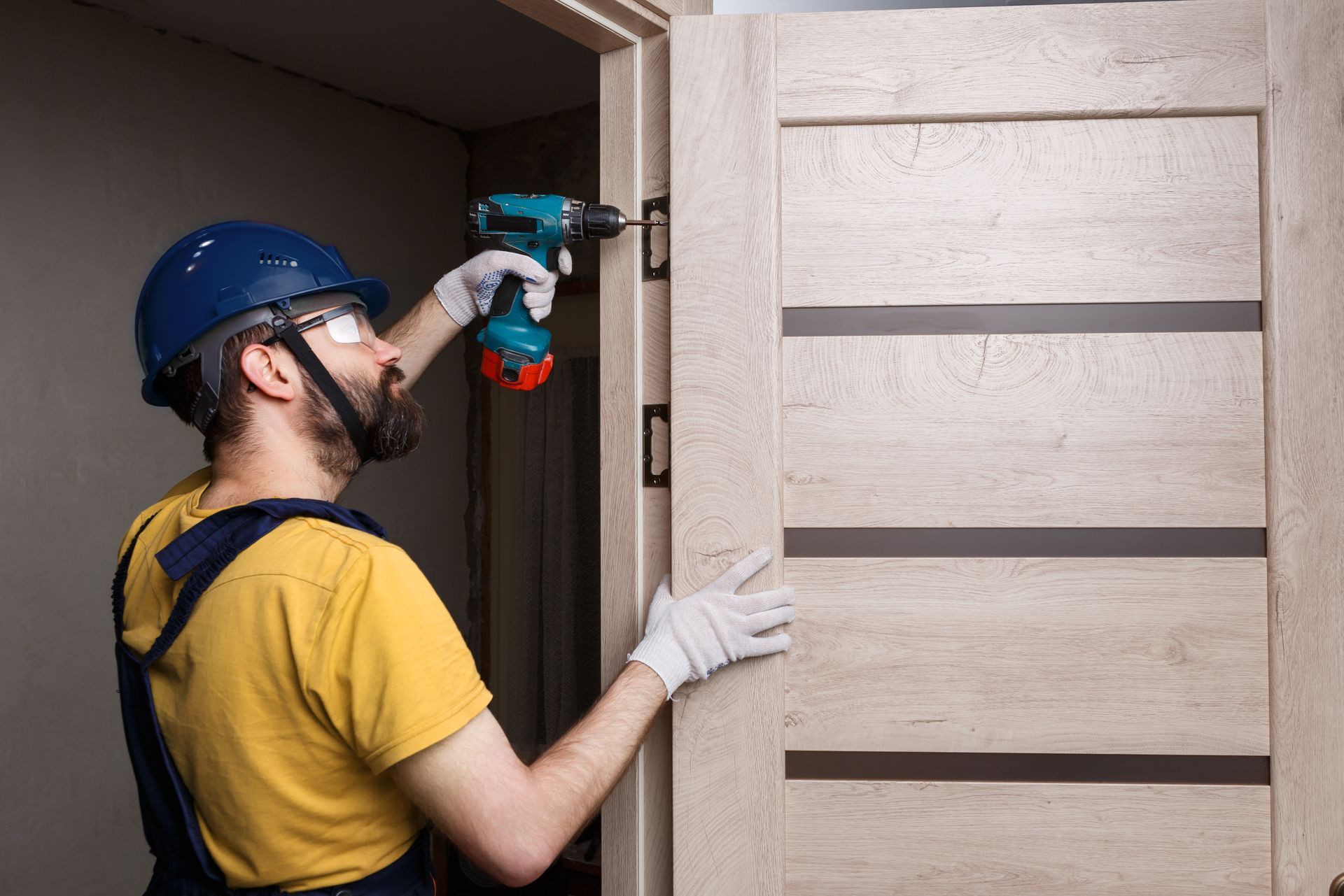 Un ouvrier perce un cadre de porte beige avec une perceuse électrique bleue, portant un casque et des gants de chantier.
