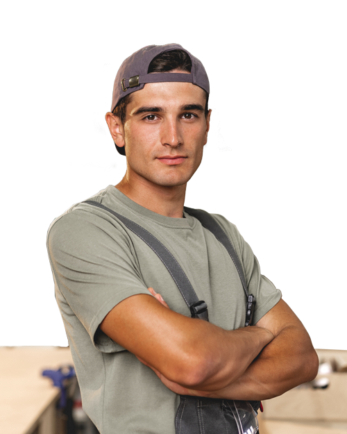 Homme les bras croisés, portant une casquette à l'envers et un t-shirt gris, se tenant devant un fond intérieur flou.