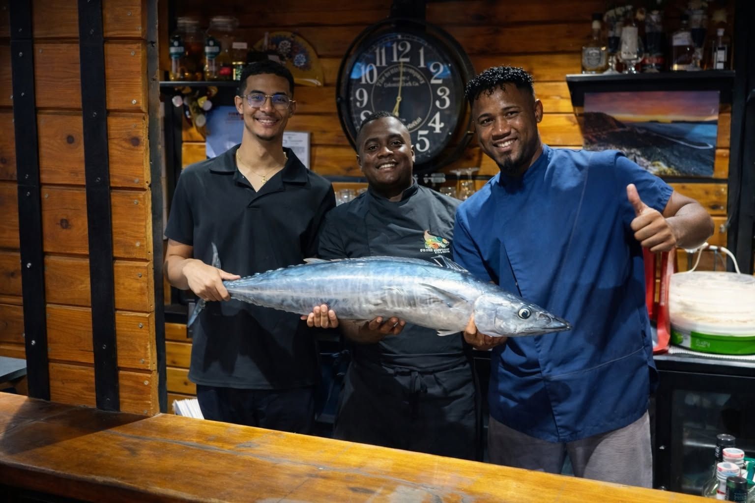 Trois personnes tiennent un gros poisson derrière un comptoir en bois, dans un restaurant.