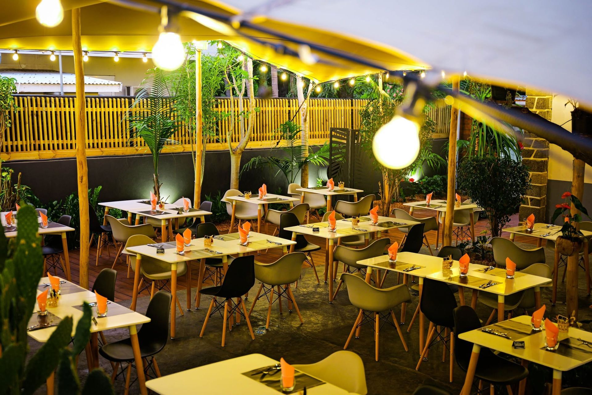 Terrasse de café en plein air avec des lumières jaunes, des tables vides et des chaises sous un auvent au crépuscule