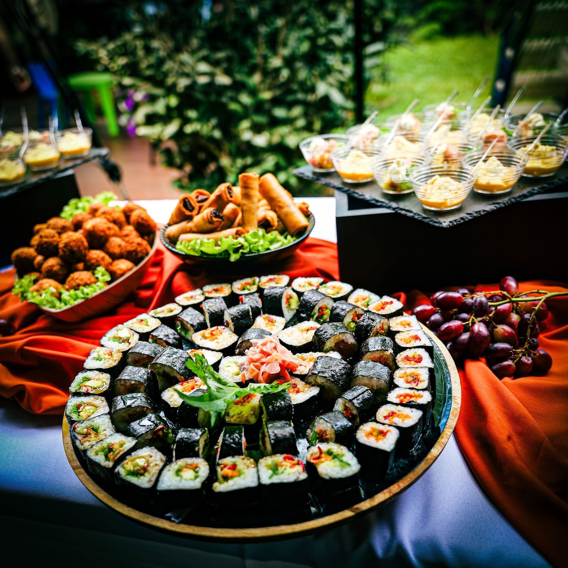 Table en extérieur avec des assortiments de sushis, des amuse-bouches frits et des coupes de dessert sur des nappes rouges