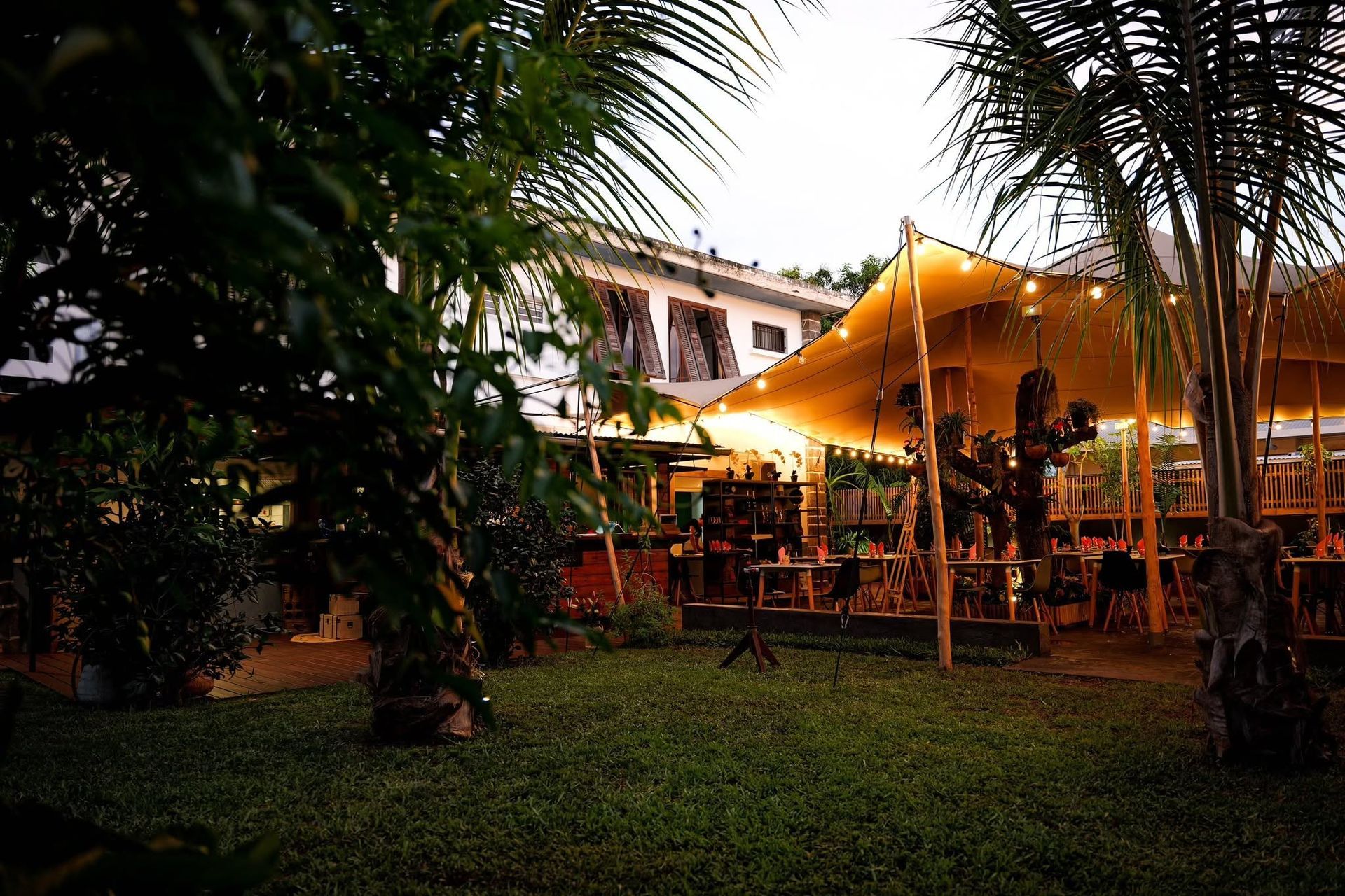 Terrasse de restaurant en plein air, avec des lumières chaleureuses, des tables et des palmiers au crépuscule