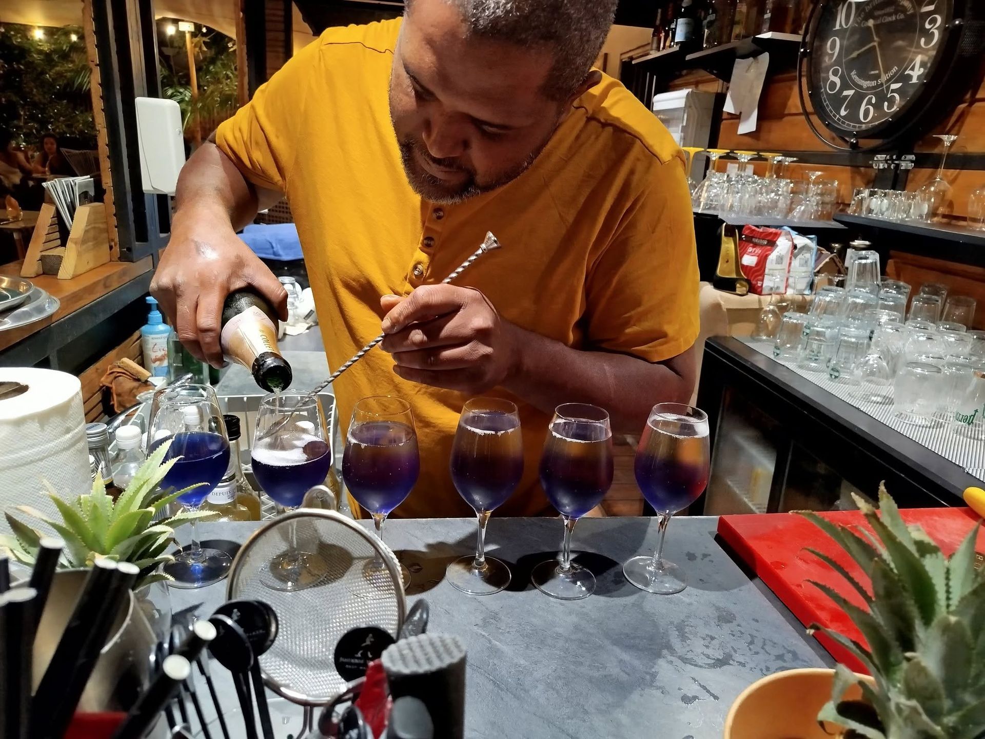Un barman verse un cocktail dans quatre verres violets posés sur le comptoir, avec de la verrerie et des objets décoratifs