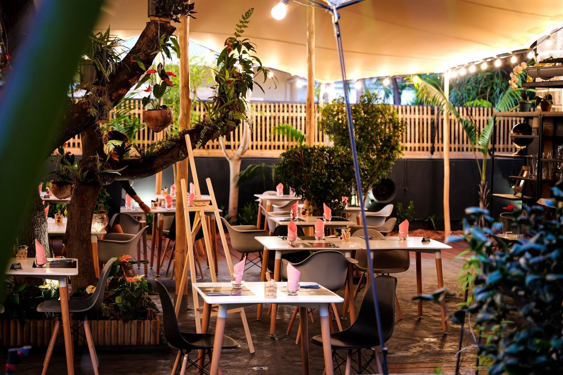 Espace de restauration extérieur convivial sous une pergola, avec des tables, des plantes en pot et un éclairage chaleureux.