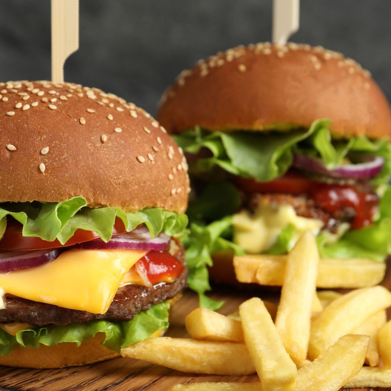 Dos hamburguesas con queso, panecillos con semillas de sésamo, lechuga, tomate y cebolla roja, con patatas fritas delante.